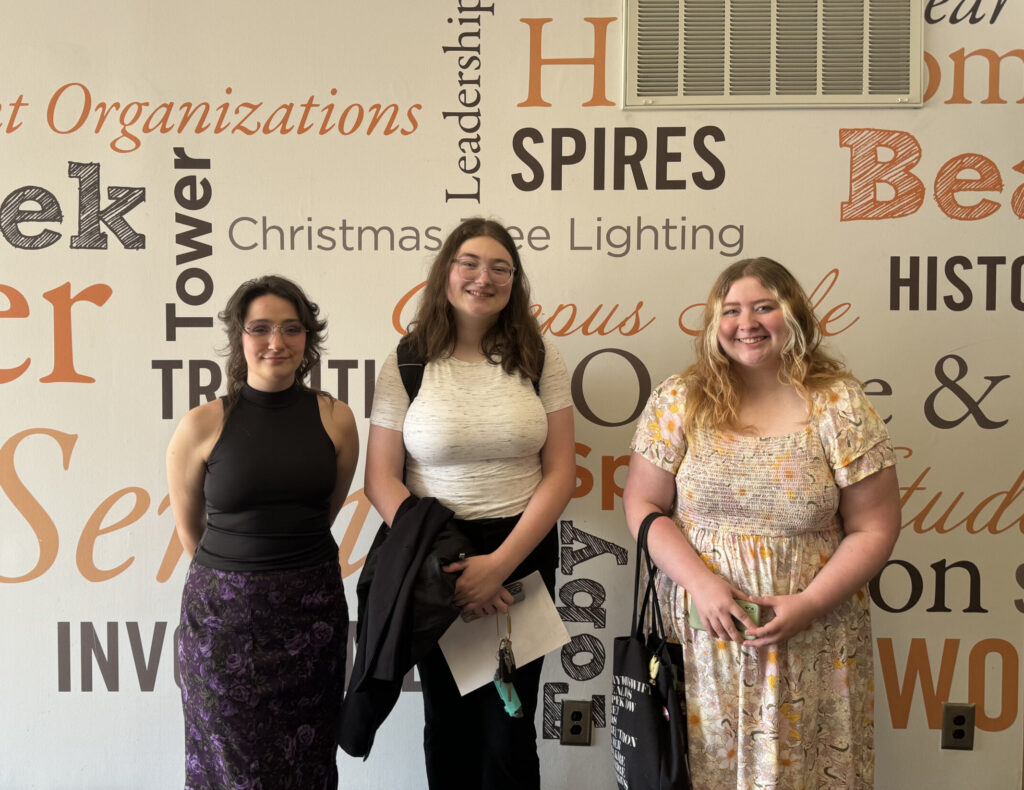 Three students standing in front of a wall with various words such as "Leadership," "Service," and "Historic" painted on it.