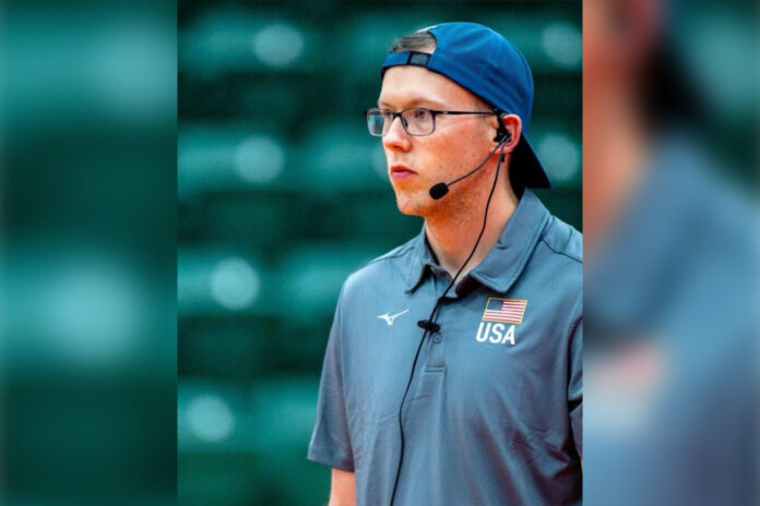 A man wears a headset, backward baseball cap and polo shirt bearing a logo with a flag and USA.