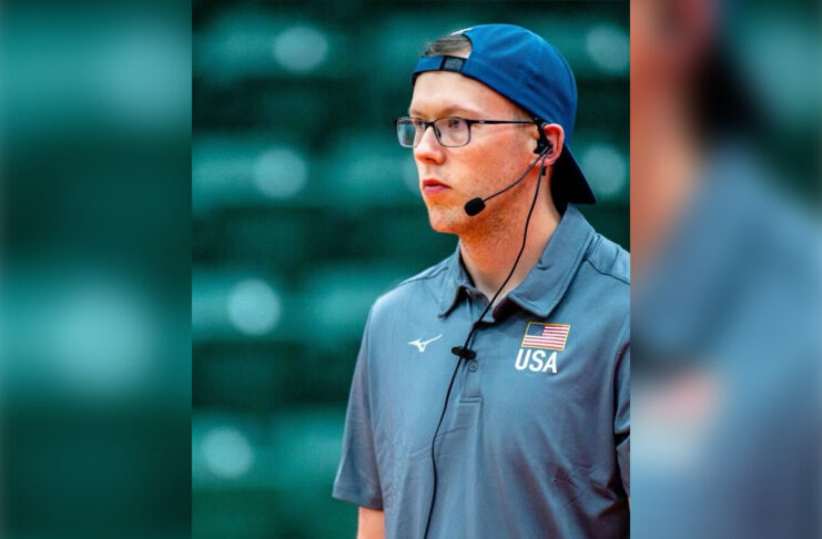 A man wears a headset, backward baseball cap and polo shirt bearing a logo with a flag and USA.