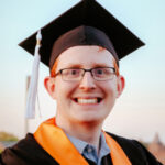 Headshot of Joshua Hynds wearing a black graduation cap and gown. He has an orange stole around his neck.