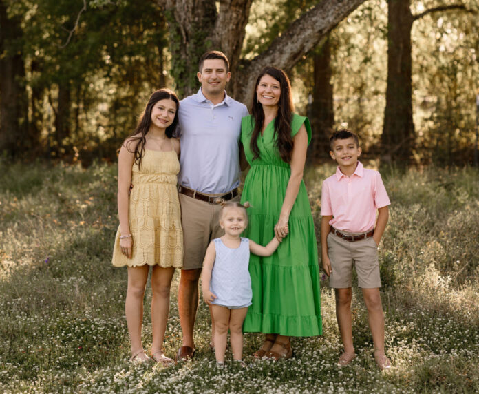 A family of five poses for a portrait in a sunlit forest setting. They are standing on a blanket of small flowers, with tall trees in the background. The group includes two adults and three children, all dressed in semi-formal attire.