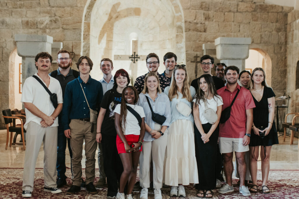 Group of 16 people posing for a photo in a stone-arched gallery, smiling at the camera.