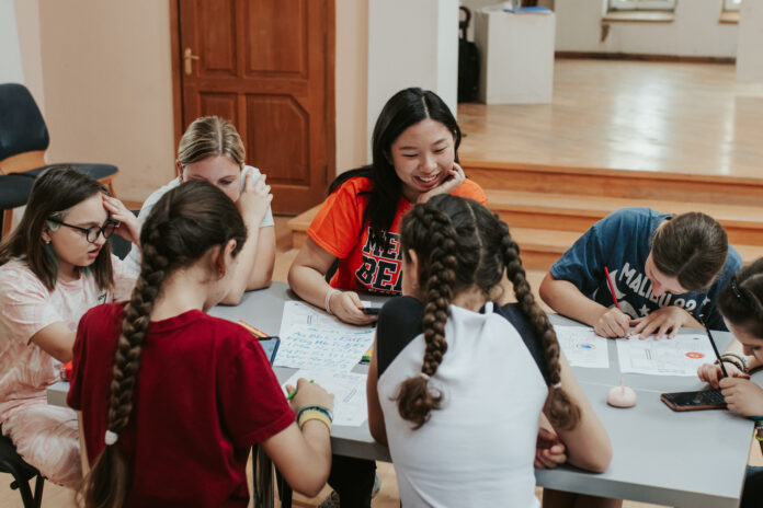 A student wearing an orange Mercer T-shirt sits at a table with children writing on papers.