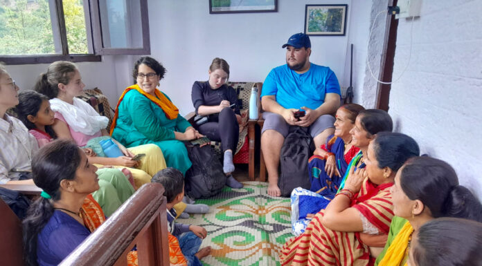 Mercer On Mission team examines effects of tourism boom in northern India Mercer students, a professor, and Uttarakhand residents sit in a circle in a room of a home. The Uttarakhand residents are wearing brightly colored traditional India clothing and sitting on the floor. The Mercer students and professor sit on a couch and chair. One student is taking notes.
