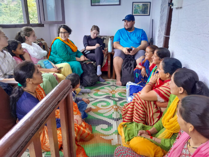 Mercer students, a professor, and Uttarakhand residents sit in a circle in a room of a home. The Uttarakhand residents are wearing brightly colored traditional India clothing and sitting on the floor. The Mercer students and professor sit on a couch and chair. One student is taking notes.