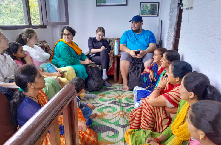 Mercer students, a professor, and Uttarakhand residents sit in a circle in a room of a home. The Uttarakhand residents are wearing brightly colored traditional India clothing and sitting on the floor. The Mercer students and professor sit on a couch and chair. One student is taking notes.