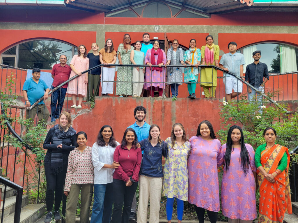 A diverse group of people stand in front of a brightly colored burnt orange building. Half the group stands on the ground and the other half stand in front of a railing on a second level. They smile for the camera.