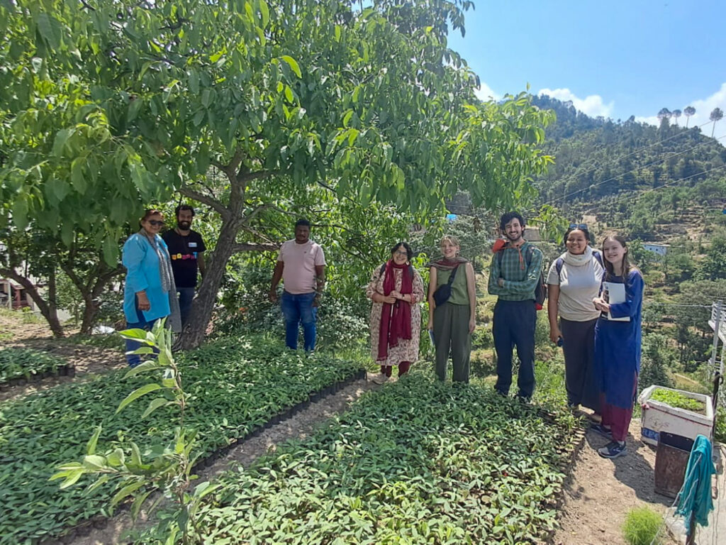 Eight people stand outside among trees and leafy vegetation. They smile for the camera.