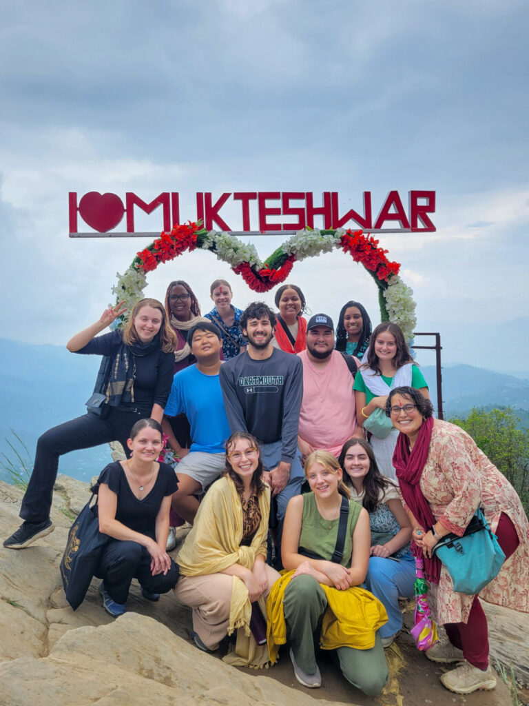 A group of 14 people pose for a picture on top of a mountain in front of a heart-shaped structure covered in red and white flowers. The words "I love Mukteshwar" are at the top of the structure.