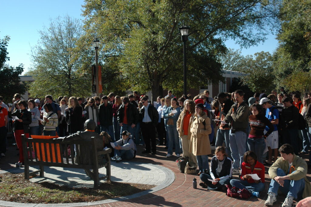 A group of people gathered around a bronze statue on a bench in a sunny park, with some attendees wearing Mercer University apparel, indicating a rally at the university.