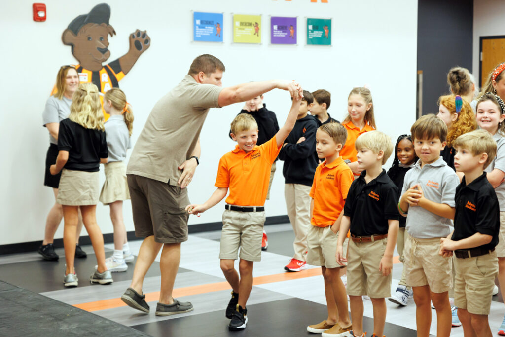 Dr. Adam Keath twirls an elementary-aged student  in a modern multi-purpose-type room. Other students and teachers are seen in the background. 