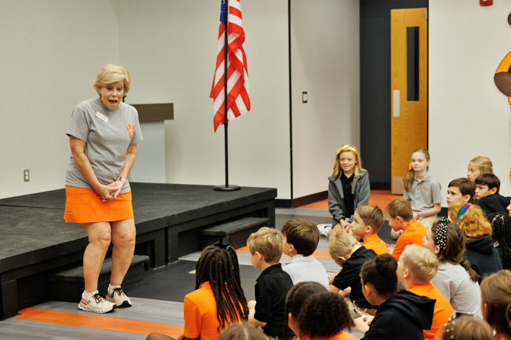 A woman talks animatedly to a group of elementary-aged students sitting on the floor. An American flag is seen in the background.