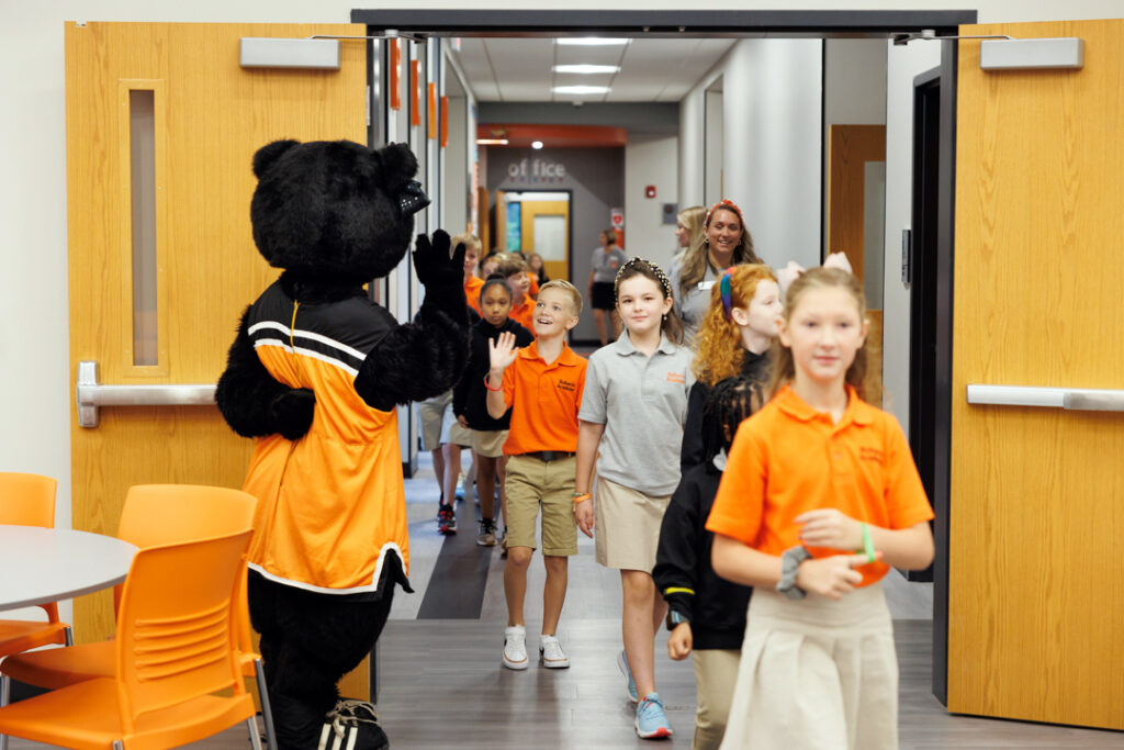 A female bear mascot waving to elementary-aged students as they walk by her in a line. 