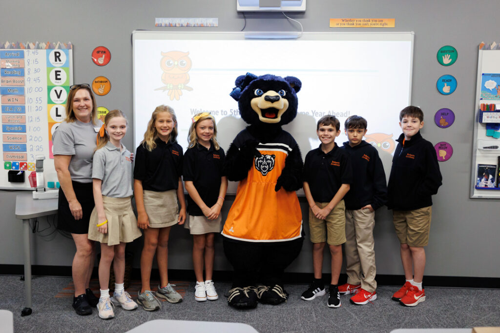 Six fifth-grade students pose for a photo standing on either side of a female bear mascot. Their teacher is on the far left. They are in a classroom setting with a whiteboard in the background. 