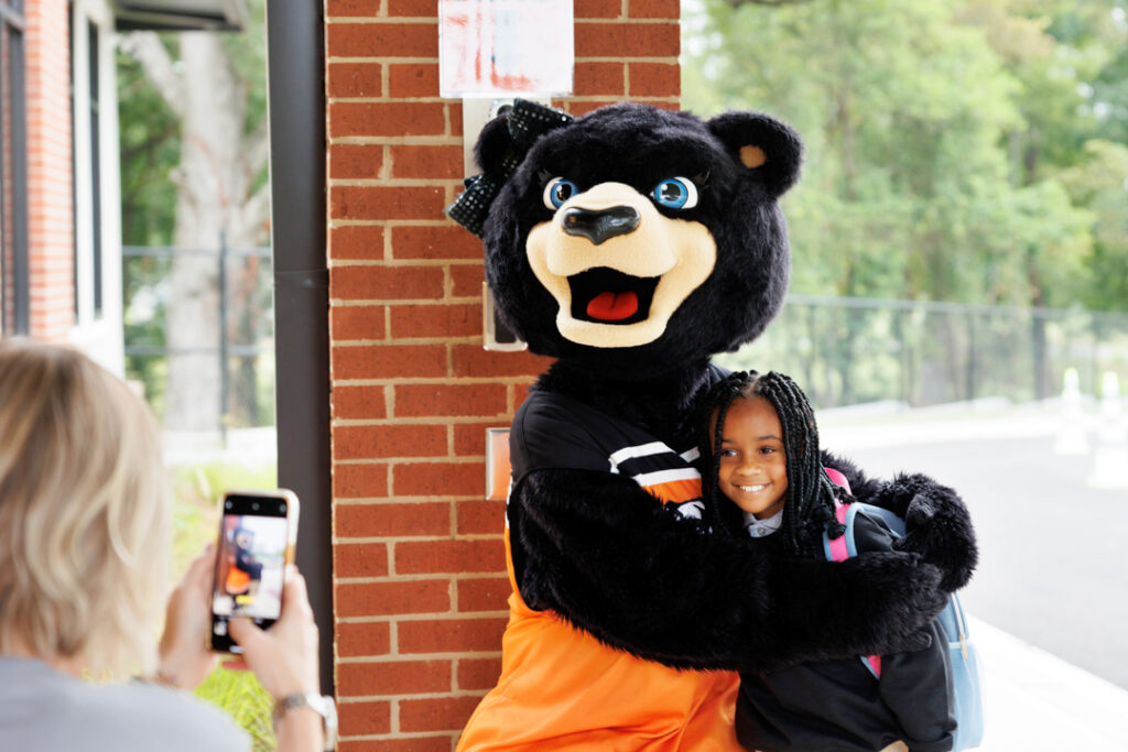 A female bear mascot hugs an elementary-aged child while a woman takes their picture with a cell phone.