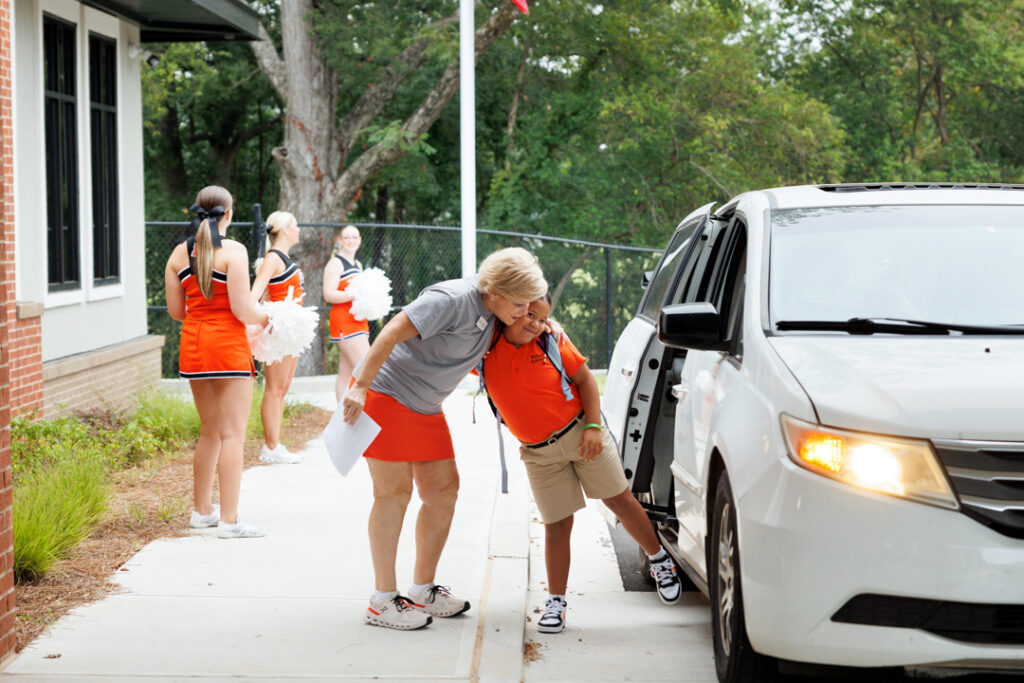 A woman hugs a child as he exits a white car. Cheerleaders are seen in the background.