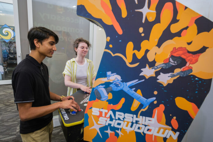 Mercer University students Rajwol Chapagain and Maret McWhorter play an arcade video game. The side of the arcade cabinet show a blue and red space ship battling each other in outer space against a blue background with yellow and orange accents. The words "Starship Showdown" are visible.