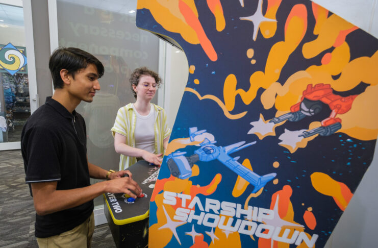 Mercer University students Rajwol Chapagain and Maret McWhorter play an arcade video game. The side of the arcade cabinet show a blue and red space ship battling each other in outer space against a blue background with yellow and orange accents. The words 