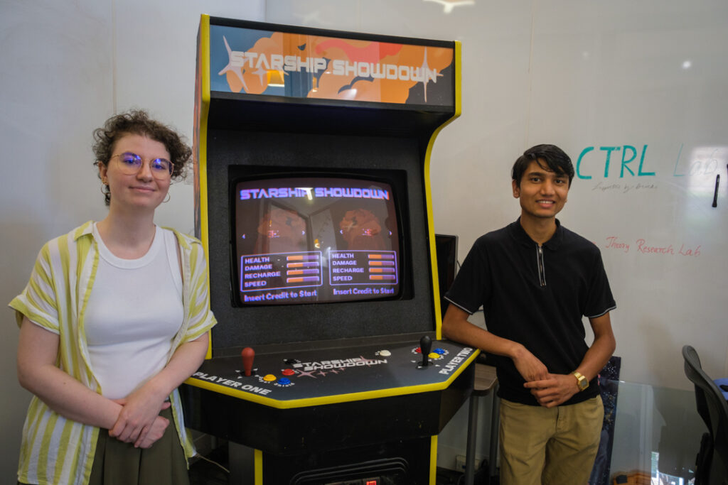 Mercer University students Maret McWhorter and Rajwol Chapagain smile while standing on either side of an arcade video game. The words "Starship Showdown" are visible on the arcade cabinet and the screen, which also shows player stats from the game.