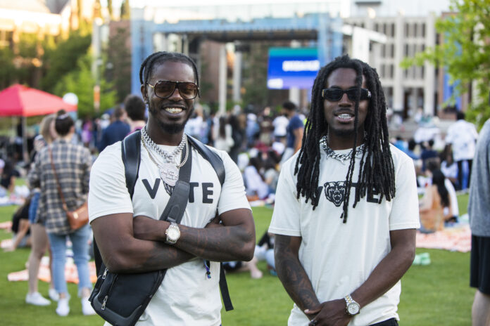 Two college students wearing Vote T-shirts stand in front of an outdoor stage.