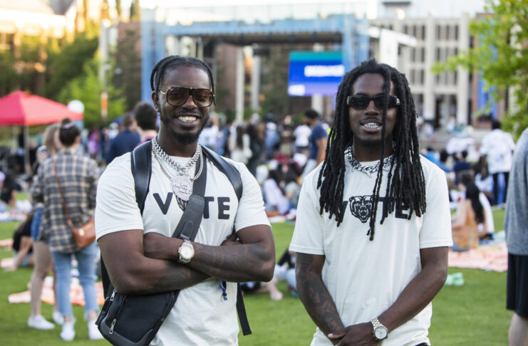 Two college students wearing Vote T-shirts stand in front of an outdoor stage.