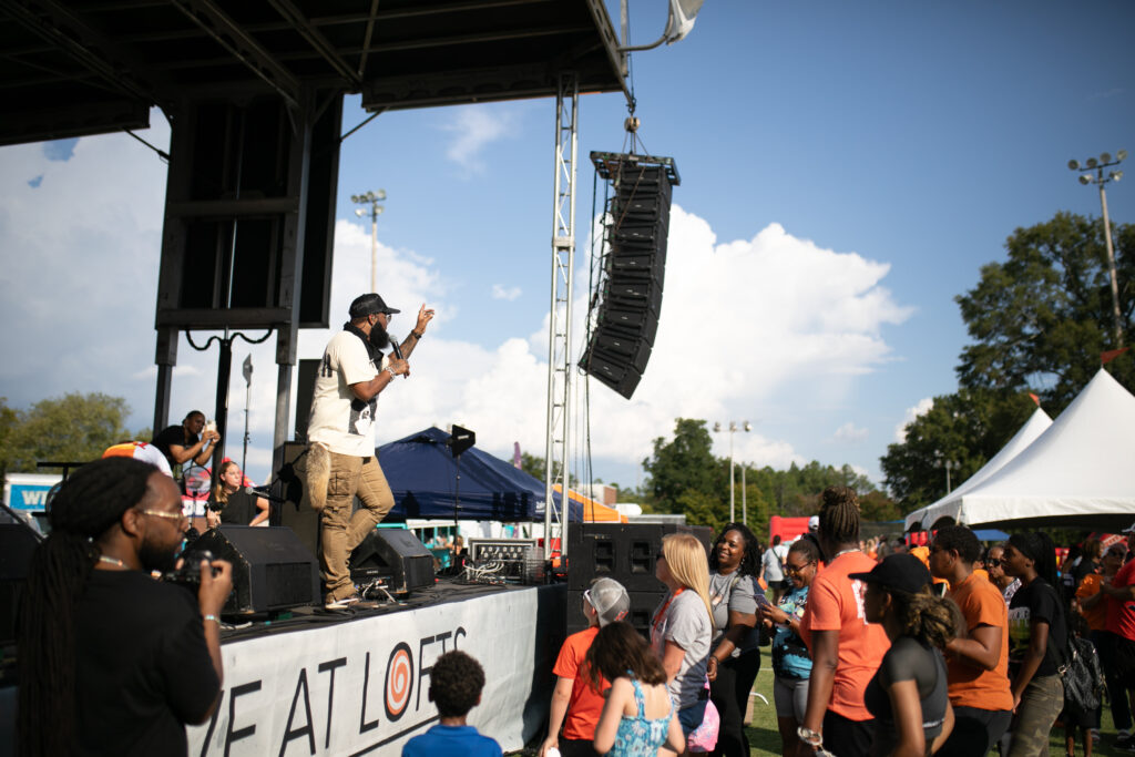 A man holding a microphone to his face performs on an outdoor stage while people look on.