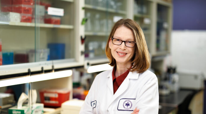 Medical school receives $176,238 grant to study effect of certain proteins on disease Dr. Pamela Cook wearing a white lab coat standing with her arms crossed in a lab.