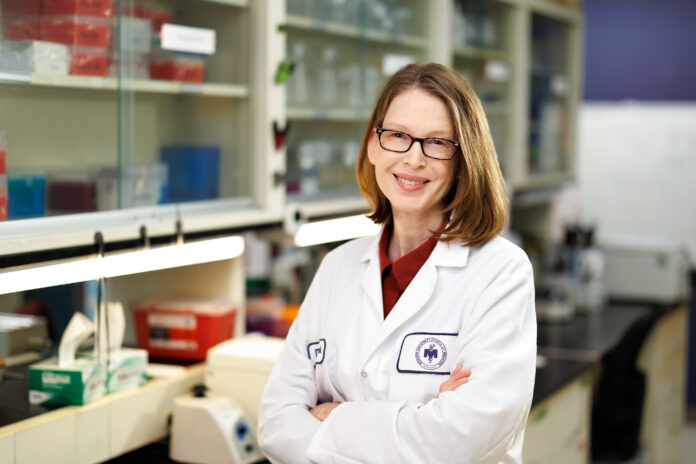 Dr. Pamela Cook wearing a white lab coat standing with her arms crossed in a lab.