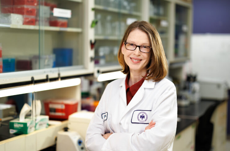 Dr. Pamela Cook wearing a white lab coat standing with her arms crossed in a lab.