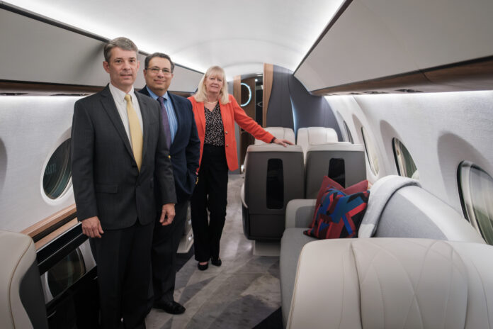 Three people in business attire stand inside a business aircraft with gray seats and walls.