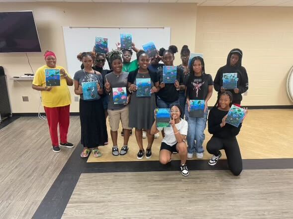 A group of people hold canvas paintings in a rec room.