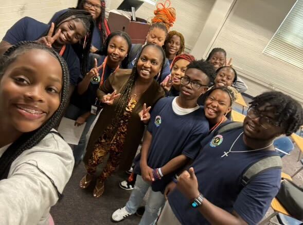 Group of happy students posing for a selfie with their mentor in a classroom, some making peace signs, wearing casual attire and school t-shirts with various badges.