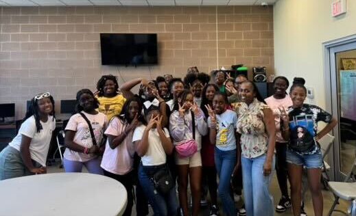 Group of young ladies posing joyfully with their mentor in a classroom with a whiteboard and audio equipment in the background.