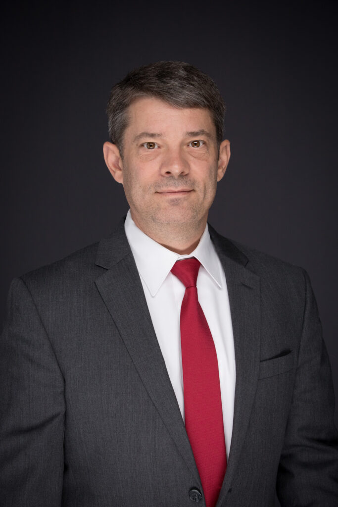 A headshot of a man wearing a red tie, white dress shirt and dark gray blazer.