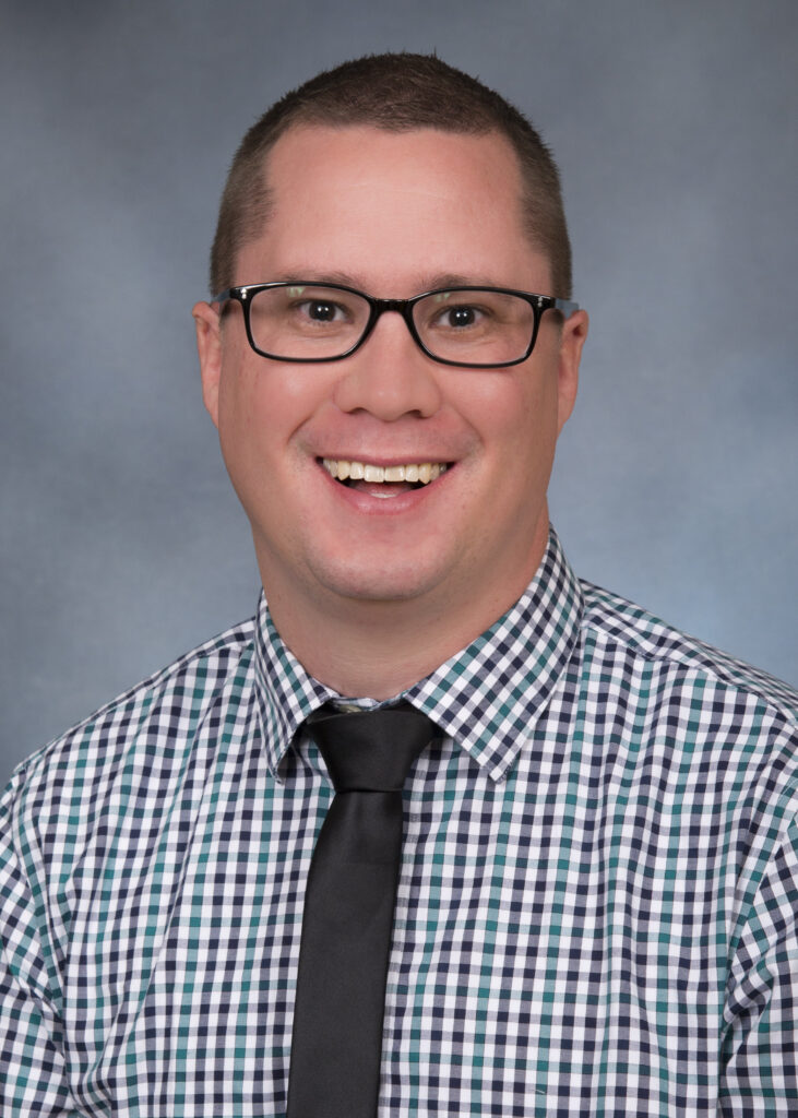 Headshot of Justin Romney wearing a checkered shirt, dark colored tie and glasses.