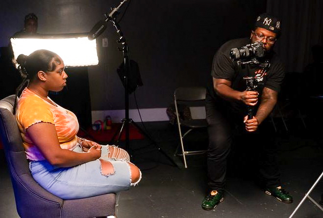 A man sits and holds a video camera pointed at a young woman who is seated and speaking to the camera.