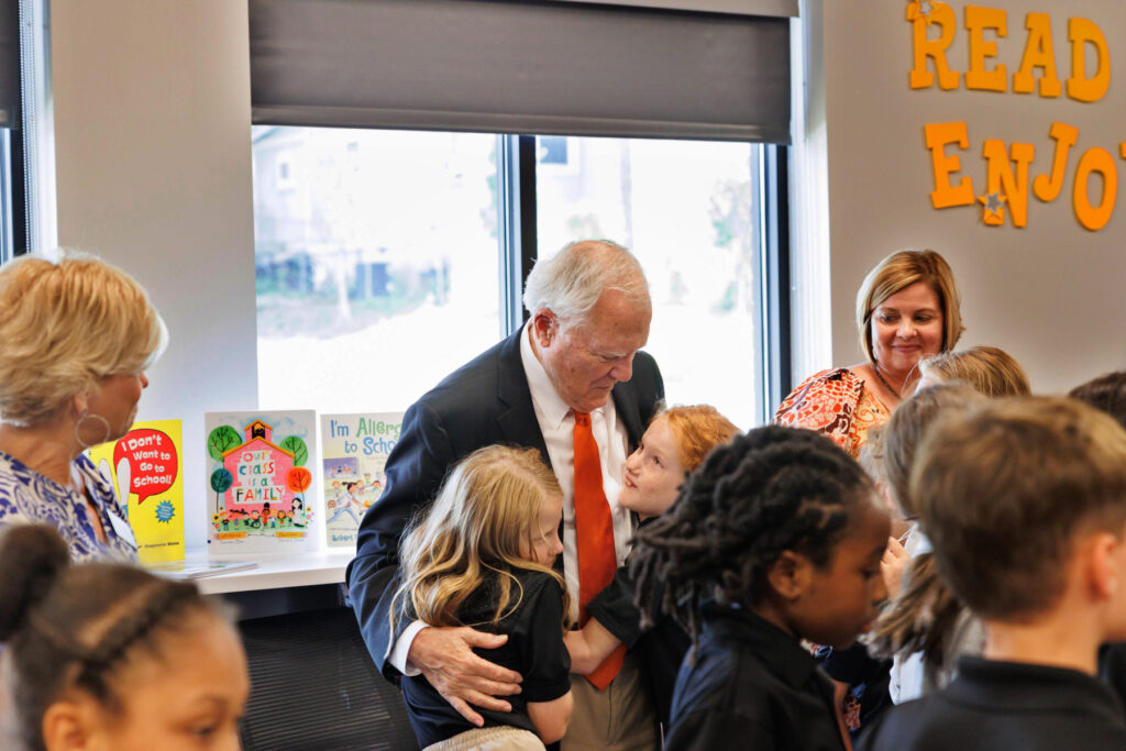 Former Georgia Gov. Nathan Deal hugs two children in a school library with other children and teachers around.