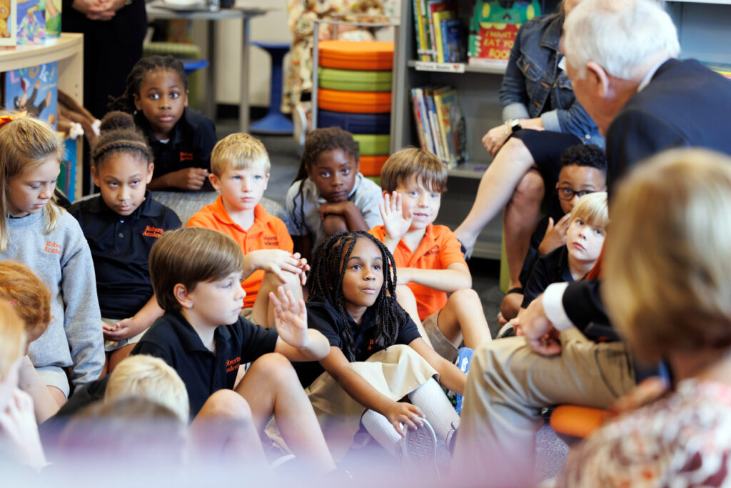 Group of children sitting on the floor in a classroom, listening attentively to an adult speaking, with one child raising their hand.