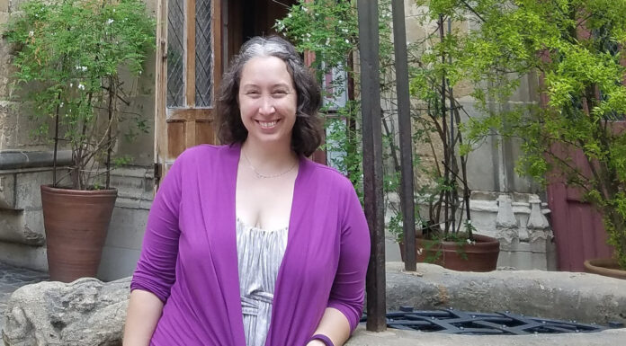 History professor to conduct landscape, garden research during yearlong fellowship A woman smiling in front of an ornate doorway with European architectural details, wearing a light gray dress and a purple cardigan.