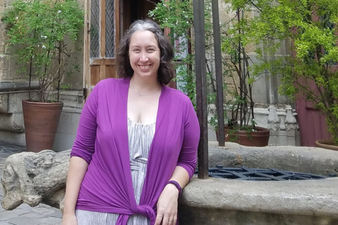 A woman smiling in front of an ornate doorway with European architectural details, wearing a light gray dress and a purple cardigan.