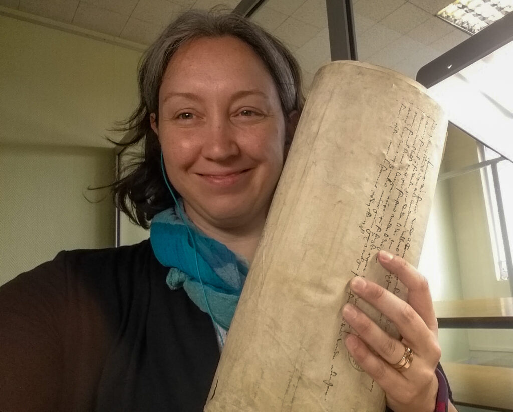 A woman smiling and holding a 1300s rolled parchment with cursive writing in a library setting.