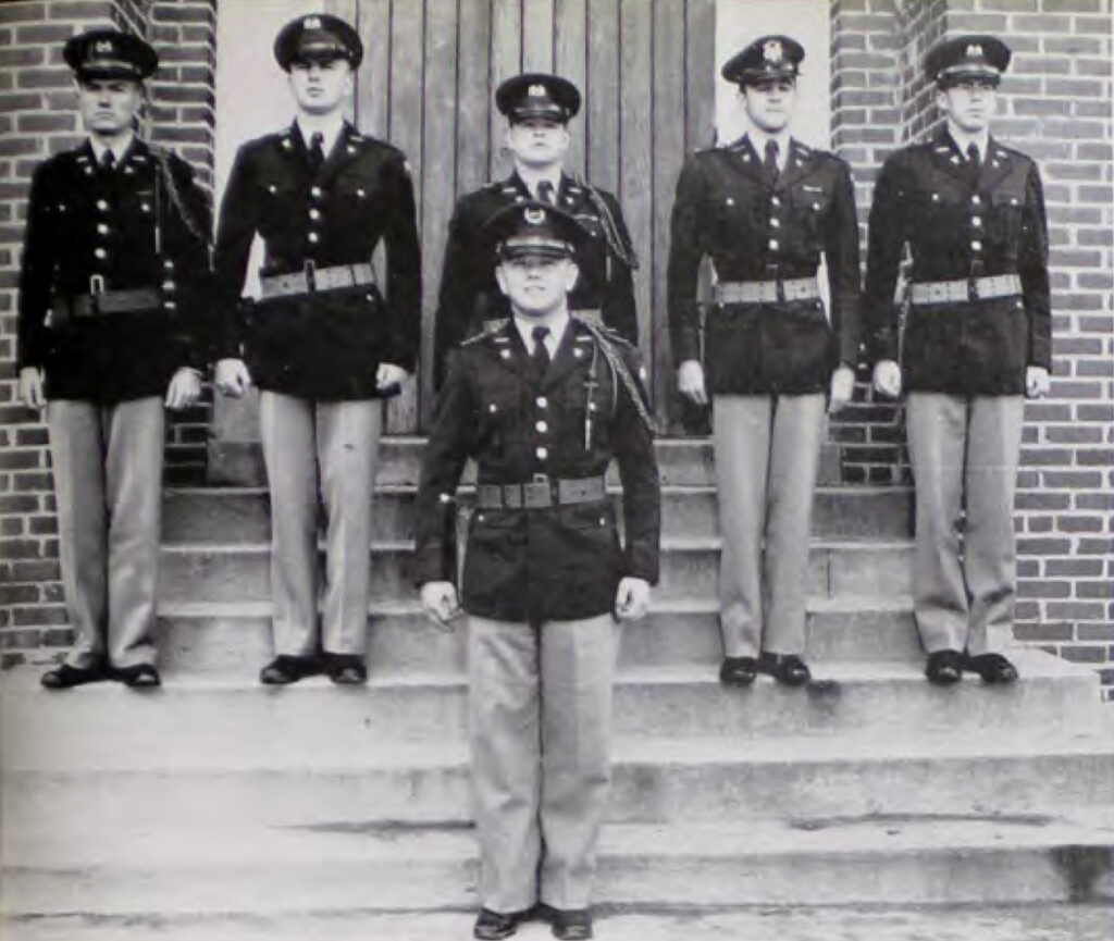 Black and white photo of six young men in military dress on the steps of a brick building.