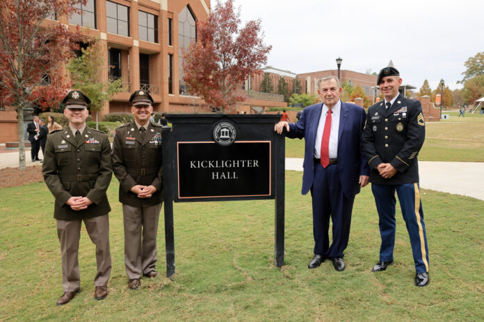 Claude Kicklighter leans his arm on an outdoor sign that says Kicklighter Hall. He is standing with three men in military unforms.