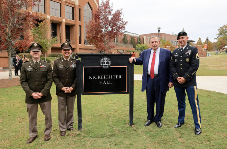 Claude Kicklighter leans his arm on an outdoor sign that says Kicklighter Hall. He is standing with three men in military unforms.