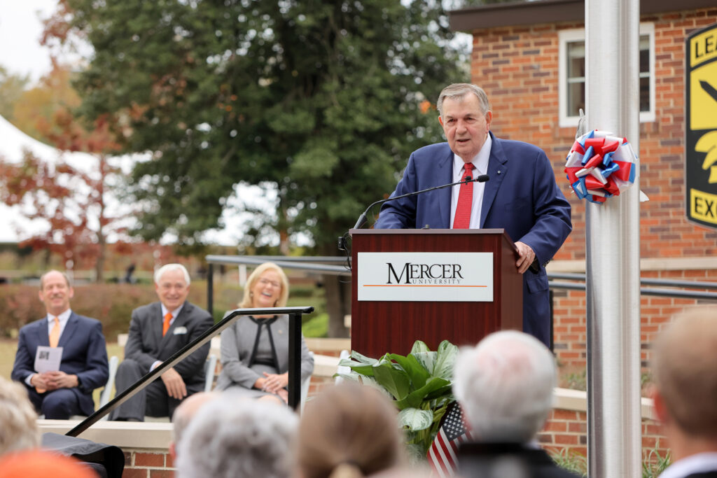 A man in a blue suit and red tie stands talking behind a podium. Others who are seated can be seen smiling in the background.