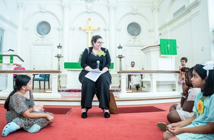 Melissa Lester sits on a chair on a red carpet in the sanctuary of a church. Children sit around her.