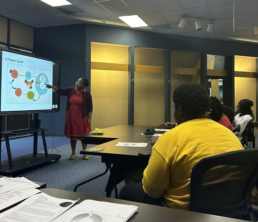 A woman stands at the front of the room and points to a PowerPoint slide on a screen, while the backs of several teens can be seen as they sit at tables and look the presentation.