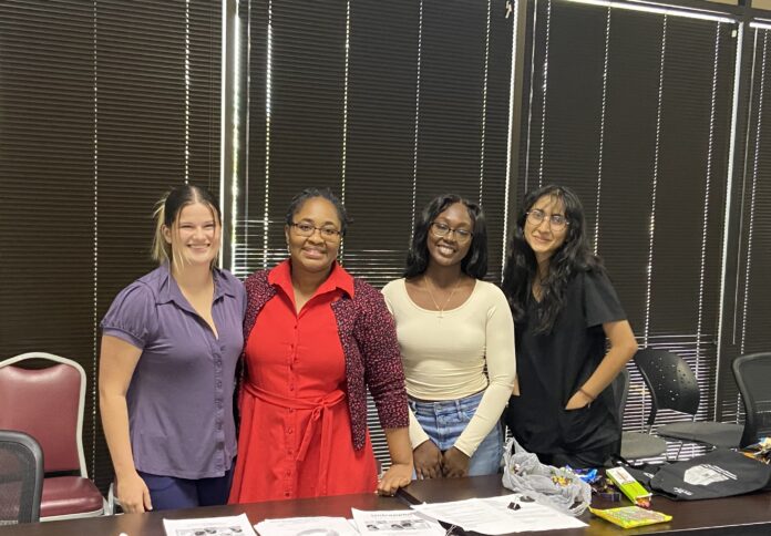 Four women stand behind a table and pose for a photo, with dark blinds on the window behind them.