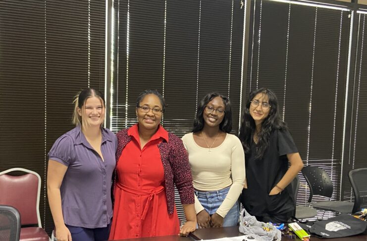 Four women stand behind a table and pose for a photo, with dark blinds on the window behind them.