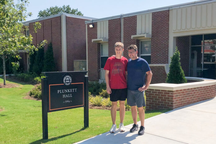 Father and son stand outside in front of a 1960s style, brick building and next to a black sign that says Plunkett Hall.